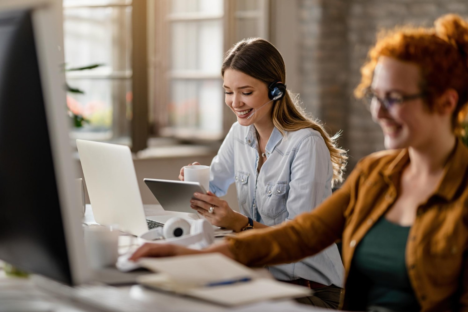 Two women working at their desks, smiling while using a tablet and computer. One is wearing a headset and holding a coffee mug, reflecting a positive and adaptable work environment. This image represents Staffing Solutions for Business Growth, illustrating how Workforce Agility creates opportunities for efficiency and engagement.